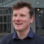 Medium young white man with dirty blond hair named James Coote, wearing blue shirt and jumper, smiling and looking slightly to one side, with some kind of modern glass fronted building in the background