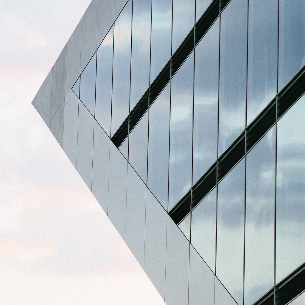 The side of a concrete and glass building with pale pink and grey clouds in the background reflecting off the windows. The building is constructed with a steep angled side such that the lower floors have a smaller footprint size than those above.